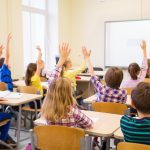 Mai mulți elevi, care stau în bănci, într-o sală de clasă, sunt cu mâinile ridicate